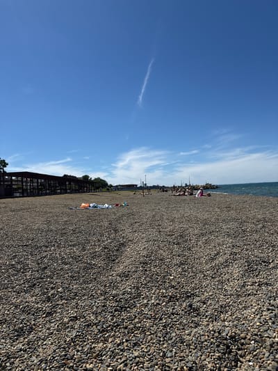 The image, correctly oriented in a landscape view, captures a bright, sunny day at a pebble beach. The dominant feature is a vast expanse of dark, small, round pebbles covering the ground, stretching from the foreground into the distance on the right side of the frame. To the left, the calm, blue waters of a sea or large lake meet the pebble shore, with subtle ripples visible on the surface. The upper portion of the image is filled with a clear, vibrant blue sky, adorned with several delicate, wispy white clouds, indicating pleasant weather.

Numerous individuals are scattered across the pebble beach, mostly concentrated closer to the water's edge in the mid-ground. They appear to be engaged in leisure activities, with some sitting or lying down on the pebbles. Several colorful items, likely beach towels, blankets, or clothing, are spread out on the beach, suggesting relaxation or sunbathing. In the far distance along the horizon, faint outlines of buildings or structures are visible. On the far left, a dark, slatted wooden structure, possibly a fence or part of a coastal boardwalk, extends parallel to the beach, partially hidden by green foliage.

The bright sunlight and clear sky suggest it is daytime, likely mid-morning or early afternoon, conducive to outdoor recreation. The scene evokes a sense of peace and leisurely enjoyment by the water. There is no visible text in the image. The setting is consistent with a coastal location, such as Taman, Russia.
