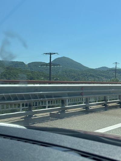 This is a medium-range, eye-level, outdoor, daytime, vertical shot taken from inside a vehicle, looking over a guardrail and towards a mountainous landscape under a clear blue sky. The foreground shows the interior of a vehicle, with a dark dashboard and the edge of the windshield. A grey metal guardrail with a red top rail separates the roadway from a green, tree-covered embankment. Beyond the guardrail, the landscape rises into rolling green hills and mountains. Several power line towers are visible on the slopes, including a prominent, tall one with a wide crossbar and insulators, and another smaller one further in the distance. The scene appears to be a highway in a rural or semi-rural area, possibly in or near Razdolnoye, Russia, given the presence of mountainous terrain and infrastructure like power lines. There is a wispy, dark smudge in the upper left portion of the sky, possibly smoke or a lens flare artifact.