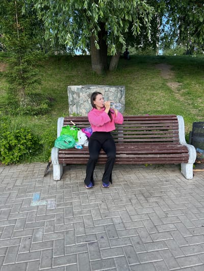The image displays an outdoor scene in Cheboksary, Russia, featuring an adult woman seated on a park bench. The woman, with long dark hair, is dressed in a pink long-sleeved cropped top, black pants, and dark athletic shoes with purple accents. She is holding a clear plastic water bottle to her mouth and appears to be drinking from it.
To her left, on the dark brown slatted bench, are multiple plastic shopping bags in green, purple, and white, some appearing to contain items. A small hand tool, possibly a trowel or shovel, with a black handle and a metallic blade, leans against the concrete support of the bench beneath the bags. The bench itself has dark brown wooden slats and light grey concrete end supports.
The ground in the immediate foreground is paved with grey interlocking bricks arranged in a herringbone pattern. Faint chalk markings, primarily in light blue and white, are visible on these paving stones. Behind the bench, a low wall constructed from irregularly shaped grey and brown stones serves as a backrest or decorative element. Beyond this wall, a grassy slope extends upwards, covered with various green bushes and trees. A large tree with weeping foliage is prominent on the right side of the background, and a smaller coniferous tree stands on the left. Two indistinct human figures are partially visible further up the grassy slope under the trees on the right. A dark metal mesh trash can is partially visible to the far right of the bench. The scene is illuminated by natural daylight.