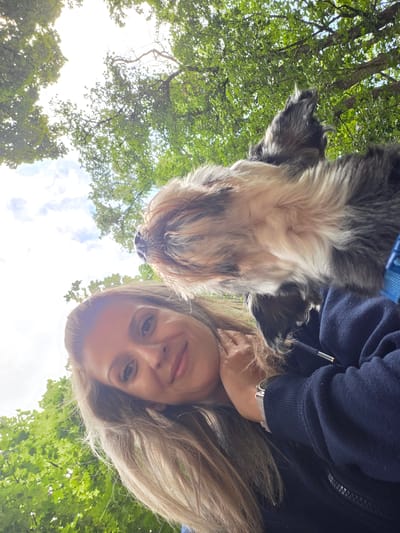 An over-the-shoulder, low-angle, outdoor selfie shows a woman and a dog. The woman is smiling, her head tilted back and her right hand resting on her neck. She has blonde hair and is wearing a navy blue hooded jacket. The dog, a shaggy, medium-sized breed, is looking upwards and appears to be wearing a blue harness. They are positioned against a backdrop of green trees and a bright sky with some clouds. The image was taken in Munich, Germany, likely in a park or wooded area.