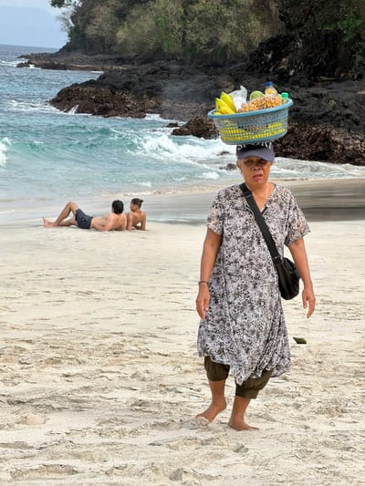 An older woman with a blue woven basket of fruit balanced on her head walks across a sandy beach in Manggis, Indonesia. The woman wears a patterned dress and dark green cropped pants, with a black bag slung over her shoulder. In the background, two individuals, a man and a woman, lie on the sand near the ocean, facing away from the camera. The ocean has turquoise water with white waves breaking against dark volcanic rocks. Trees and vegetation cover a hillside behind the rocks. The sand is light brown and textured with ripples and footprints.