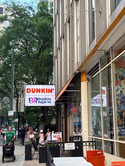 The image shows a street-level view of a Dunkin' and Baskin-Robbins combination store in an urban environment, likely Chicago, United States of America, based on the architecture and street signs. The store has a large, prominent sign featuring the Dunkin' logo in orange and the Baskin-Robbins logo in pink. The storefront features large glass windows and an awning. Outside the store, on the sidewalk, several people are visible. A woman in a green shirt pushes a stroller. Another woman in a pink dress is walking, holding a phone and a purse. Two young girls, one in a pink dress and the other in a white dress, are standing near a menu board displaying a "$6 Meal Deal". The street itself shows traffic and other pedestrians in the background. Lush green trees are present on the left side of the image, providing a contrast to the urban buildings. The overall scene depicts a busy commercial street with a focus on the food establishment and its customers.