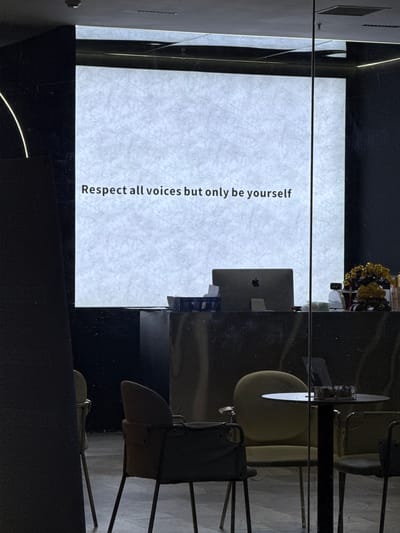 The image shows a framed piece of paper with the text "Respect all voices but only be yourself" displayed vertically. It is positioned against a backdrop of modern office furniture, including a desk with a computer, and chairs. There is also a decorative floral arrangement on the desk. The scene is reflective, suggesting a glass or polished surface in the foreground. Without additional information, it is impossible to pinpoint the precise location within the Chaoyang District, China.