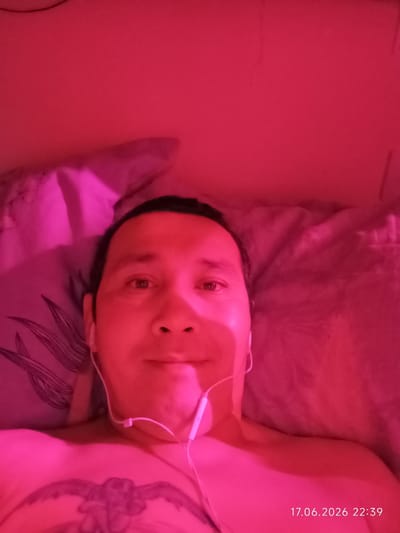 A close-up selfie shows a person lying down, illuminated by pink light. The person's head is angled slightly towards the camera, and they are wearing white earbuds. Their chest, partially visible, has a tattoo. The background appears to be a bed with patterned pillows. The image is dated "17.06.2026 22:39," suggesting it was taken in the evening. The setting is likely a bedroom in Nyurba, Russia.