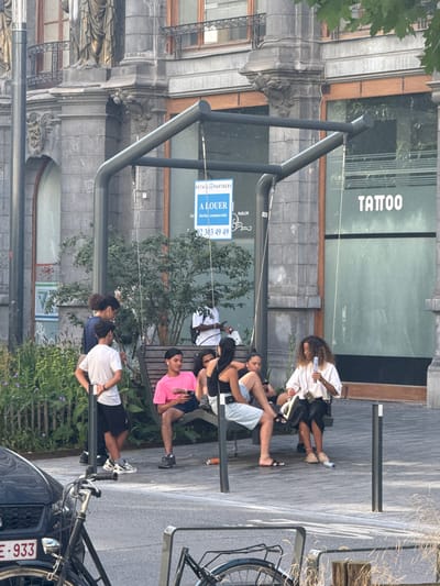 The image depicts a street scene in Brussels, Belgium, with several people sitting on a bench. The setting appears to be outdoors, possibly in a commercial area given the presence of a building with "TATTOO PARLOR" signage and a "A LOUER" (For Rent) sign on the facade. The time of day seems to be daytime, with bright lighting suggesting fair weather.
The individuals on the bench are casually dressed, and their postures suggest relaxation. One person, a woman with curly hair, is holding a bottle. Another person is wearing a pink t-shirt and dark shorts, while others are in white t-shirts and dark shorts or skirts. There's a bicycle parked nearby, a common sight in urban European settings. The architecture of the building in the background shows ornate details, hinting at an older part of the city.
The visible text includes "TATTOO PARLOR", indicating the nature of a business located there. The "A LOUER" sign suggests a commercial space is available for lease. Additionally, a phone number "02 303 49 49" is visible on the rental sign. The license plate of a car in the foreground shows "E-933". The overall mood seems relaxed and ordinary, capturing a moment of everyday life in the city.