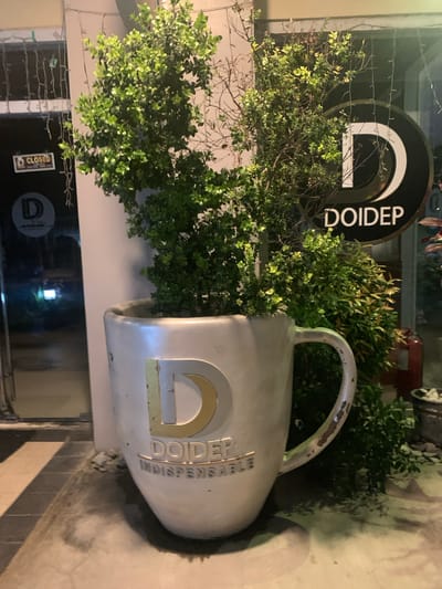 The image captures an outdoor scene in Angeles, Philippines, likely at dusk or during the evening, given the artificial lighting. The primary subject is a large, mug-shaped planter made of a distressed, light-colored material. This planter is overflowing with lush green foliage, suggesting a well-maintained garden or decorative element outside a business.
Prominently displayed on the planter is the logo "DOIDEP INDISPENSABLE" in gold lettering, with a stylized "D" also rendered in gold. To the left of the planter, a circular black sign with a white "D" and the text "DOIDEP" is visible, partially obscured by the foliage. This suggests that the planter is a decorative feature for a business named DOIDEP.
In the lower left portion of the image, a storefront window is visible. A "CLOSED" sign is attached to the glass. The reflection in the window shows a dark interior, further reinforcing the idea that it is an establishment. The overall setting appears to be a commercial area, with the planter serving as an eye-catching display. The lighting creates a slightly dramatic ambiance, highlighting the textures of the planter and the vibrant green of the plants. There are no people visible in the frame.