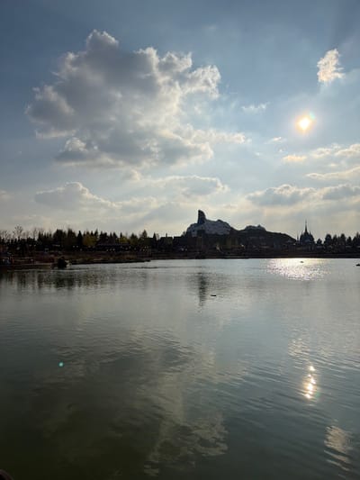 The image depicts a wide view across a large body of water, likely Lake Disney, within Disneyland Paris in Chessy, France. The scene is illuminated by the sun, positioned in the upper right quadrant of the sky, casting a bright glare on the rippled water surface towards the right. The sky is partially cloudy, featuring scattered cumulus clouds against a light blue expanse. The distant shoreline in the background is lined with evergreen trees and various themed structures. A prominent artificial mountain, identifiable as Big Thunder Mountain, stands in the center-right background, partially covered in white and featuring a distinct pointed peak. To its right, a dark, spire-like structure resembling a castle is visible. On the far left, a small pier or platform extends into the water, with several indistinct human figures seated upon it. A small, dark, oblong object floats on the water's surface in the mid-distance. The overall lighting suggests a bright, possibly late afternoon or early morning, outdoor environment.