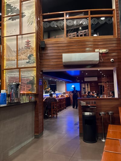 The image depicts the interior of a Japanese restaurant, likely in Magdalena del Mar, Peru, characterized by its extensive use of dark wood paneling and ambient lighting. In the foreground, a bar area is visible with a stainless steel coffee maker, a blue thermos, glasses, and bottles on the counter. To the right, a man is seated at the bar, facing away from the camera. Beyond the bar, an open entryway reveals the main dining area. Here, a staff member in dark attire is walking past a sushi bar where patrons are seated. A large screen displays the "Paramount" logo. The restaurant features tiered seating and decorative elements reminiscent of Japanese art, such as paintings of landscapes and pagodas on the upper levels. The overall atmosphere appears to be upscale and contemporary, with a clear emphasis on a Japanese theme.