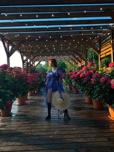 In Verkhneye Uchdere, Russia, a woman with red hair stands on a wooden deck beneath a pergola covered with string lights. She wears a flowing blue and purple sheer dress, a black belt, and black boots. In her hands, she holds a wide-brimmed straw hat. The deck is lined with large potted hydrangeas in various shades of pink and purple. The wooden planks of the deck appear wet, reflecting the low-angled sunlight. Green foliage is visible in the background, suggesting an outdoor setting.