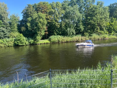 An eye-level shot shows a boat with a blue hull and white top moving to the right on a wide, dark river. The river is bordered by a lush, green treeline on the left bank, with dense trees and some shorter vegetation along the edge. A wire fence in the foreground with tall green grass growing through it partially obstructs the view of the river and the vegetation. The sky is clear and blue. The location is the Havel canal in Teltow, Germany.
