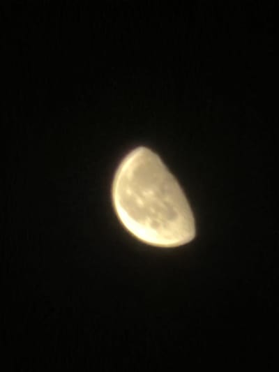 The image displays a celestial body, identifiable as the Moon, captured against a uniformly dark, black background, indicative of a nighttime sky. The Moon is illuminated, appearing in a waxing gibbous phase, with approximately two-thirds of its disk visible and brightly lit. Its color is a warm yellow-gold. Visible features on the lunar surface are indistinct due to a slight blur or soft focus, but darker areas, likely maria, are discernible within the illuminated portion. The edges of the Moon show a subtle glow or chromatic aberration. There are no other celestial objects, atmospheric phenomena, or terrestrial elements visible in the frame. The image does not contain any people, subjects, objects, or environmental details that would indicate a specific terrestrial location, including the city of New Delhi, India.