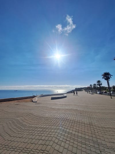 This image depicts a seaside promenade on a bright, sunny day. The sky is a vibrant blue with a few wispy clouds, and the sun shines intensely, casting a bright glare on the water. The sea stretches out to the horizon, calm and reflective, with several small boats visible.
The promenade itself is paved with rectangular brown bricks laid in a diamond pattern. It curves gently along the coastline. Along the edge of the promenade, facing the sea, is a low, curved wall.
In the distance, on the landward side of the promenade, a line of palm trees and other greenery can be seen, suggesting a warm, coastal climate. A few people are walking along the promenade, appearing as small silhouettes against the bright background. A single car is visible near the palm trees.
The lighting indicates it is daytime, likely mid-morning or early afternoon, given the angle of the sun. The overall impression is one of a pleasant, leisurely day at the beach or seaside resort. There is no visible text in the image.