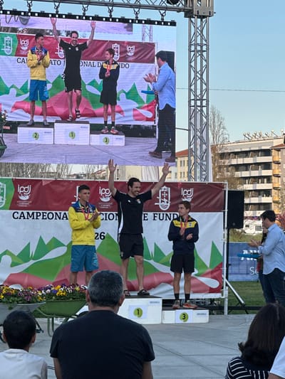 This image depicts an outdoor awards ceremony taking place in Mirandela, Portugal. On a central stage, three male athletes are positioned on a three-tiered podium, numbered "1" (center), "2" (left), and "3" (right), with "MIRANDELA" visible on the podium steps. A large LED screen directly above the stage mirrors the live scene below.
The athlete on the first-place step (center) is a man with dark hair and a beard, dressed in a black short-sleeved top, black shorts, and black sandals. He has both arms raised in a celebratory gesture and is smiling, wearing a medal around his neck. The athlete on the second-place step (viewer's left) is a young man in a yellow long-sleeved jacket, blue shorts, and light-colored athletic shoes. He holds a medal in front of him. The athlete on the third-place step (viewer's right) is a young man in a dark blue and yellow long-sleeved top, black shorts, and orange and grey athletic shoes, also holding a medal.
To the right of the podium, an official, a man in a light blue collared shirt and dark trousers, stands clapping, with a blue object in his left hand. A large banner behind the podium displays the text "CAMPEONATO NACIONAL DE FUNDO," along with the logos of IPDJ and FPC, and stylized green and white mountain graphics. A black speaker is visible to the right of the stage. To the left of the podium, a green wheelbarrow holds a collection of colorful potted plants.
In the immediate foreground, the back of a man's head and shoulders, wearing a black t-shirt, is visible, along with parts of other spectators to his left and right. The background features multi-story urban buildings and some bare trees under a clear, bright blue sky, indicating daylight conditions.