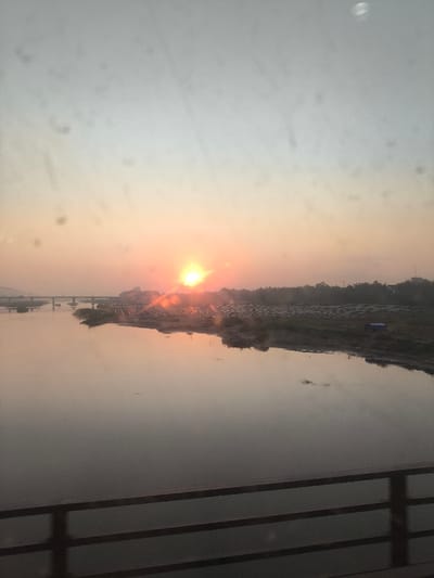 The image displays a sunrise or sunset over a wide river in Sơn Tịnh, Vietnam, observed from an elevated perspective, likely a bridge, with a multi-bar dark railing visible in the immediate foreground. The sky transitions from a light blue-gray at the top to a prominent orange and pink hue near the horizon. A bright, intensely glowing orange-yellow sun is situated slightly left of center on the horizon, emitting distinct rays and reflecting off the water. The calm river, occupying the mid-ground, reflects the warm colors of the sky and sun. In the far distance to the left, a multi-span bridge crosses the river. The right bank of the river features an expansive, flat area containing numerous organized rows of low-lying structures or coverings that reflect the sunlight; a small, blue tarp-covered object is present on this bank further to the right. A dense line of dark vegetation or trees forms the distant horizon beyond this developed area. No individuals, direct actions, or interactions are visible within the scene. The image contains various dust particles or smudges, particularly in the upper sky region.