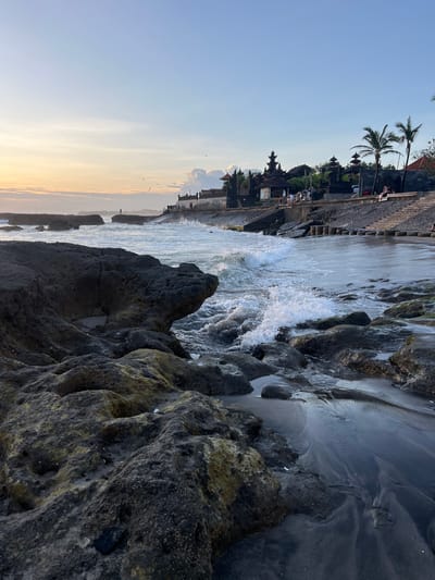 This is an eye-level landscape photograph, rotated 90 degrees clockwise, taken in Canggu, Indonesia, featuring a rocky shoreline, ocean waves, and a coastal structure.

The foreground is dominated by large, dark, and weathered rocks with visible textural details. In the middle ground, ocean waves crash against the rocks, creating white foam and turbulent water. A sandy, dark-colored beach area is visible near the rocks.

In the background, a structure stands on a raised platform, with visible palm trees and architectural details suggesting a temple or significant building. The sky is gradient, transitioning from a pale blue near the top to a warm, orange hue closer to the horizon. A few birds are visible in the sky. Several people can be seen standing along the barrier.
