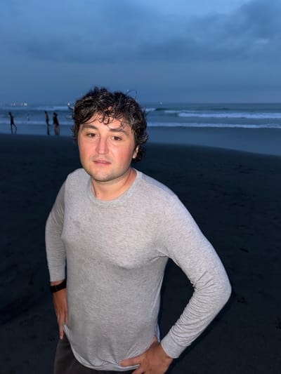 A man stands on a dark sandy beach with ocean waves in the background under an overcast sky, likely at dusk or in the evening. The man is wearing a light gray, long-sleeved shirt and dark pants. He is looking at the camera with a slight smile. His hair is dark and wet, possibly from the sea. Based on the lighting, beach appearance, and vegetation in the background, the location is likely Canggu, Indonesia.