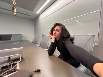 The image depicts a modern office conference room, stated to be located in Garston, United Kingdom. The room features light grey walls and a white ceiling with linear recessed lighting and a gold-colored cylindrical decorative fixture. A single female individual, with dark wavy hair, is positioned in the foreground, taking a selfie. She is wearing a black long-sleeved ribbed top and dark lower garments, with her left hand resting against her jawline. Her expression is neutral as she looks directly at the camera. In the middle ground, a long, dark grey conference table extends from the foreground to the background, surrounded by several light grey office chairs with mesh backs. On the table in the foreground, a white Cisco device, various black and grey cables, and a dark grey rectangular Belkin device are visible. A large glass partition forms part of the back wall, reflecting the overhead lights. Behind the table on the left, a dark credenza or cabinet with a black top is present. The overall environment appears clean, organized, and professionally corporate.