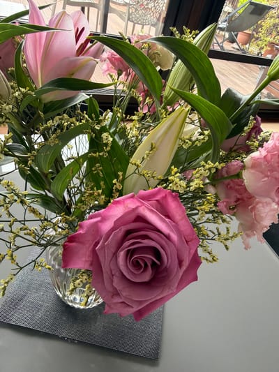 A bouquet of flowers, primarily pink roses and lilies, is arranged in a clear glass vase. The bouquet also includes green leaves and small yellow flowers. The vase rests on a dark gray, textured surface. A window with a view of trees and a building exterior is visible in the background. It is not possible to verify the location of the city of Champs-sur-Marne, France from the context.