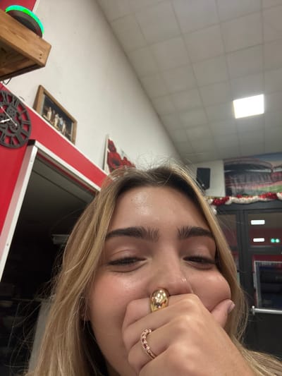 The image depicts a young individual with blonde hair posing indoors, covering their nose and mouth with their right hand in a playful or bashful gesture. The person's eyes are closed or squinted in a smile. Two rings are visible on the right hand: a large, polished gold dome-shaped ring on the thumb and a rose gold band with small red stones on the ring finger.
The background reveals a commercial interior with white walls, a red horizontal stripe/panel, and a white ceiling composed of square acoustic tiles with a rectangular fluorescent light fixture. On the left, a wooden shelf holds a green glowing circular object. Below the shelf, a dark, ornate clock with Roman numerals is mounted on the wall. Further right, a framed picture and a partial view of an artwork with red elements are visible. On the right side of the frame, a black wall-mounted speaker is present, and red and white garland decorates what appears to be a doorway or window. Beyond this, a blurred image of a stadium's seating area is discernable.
Based on the distinct stadium backdrop and the specified location, this scene is situated within or adjacent to a venue associated with the **Parc Olympique Lyonnais (Groupama Stadium)** in Décines-Charpieu, France, likely a concession stand, restaurant, or lounge area within the stadium complex or nearby.