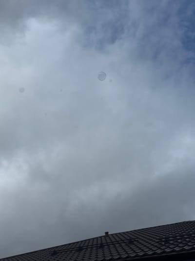 The scene depicts an upward-angled view towards an overcast sky over Krasnooktiabrskiy, Russia. The upper approximately 80% of the image is dominated by a cloudy sky, showing varying shades of grey and white clouds with occasional hints of lighter blue. Numerous translucent, spherical objects, consistent with soap bubbles, are visible suspended in the sky; one larger, distinct bubble is located above the center-right, with several smaller, fainter bubbles dispersed throughout the mid-ground and left side of the sky. The bottom approximately 20% of the image displays a section of a dark brown or dark grey tiled roof, featuring overlapping, textured tiles. A dark, cylindrical vent or pipe protrudes from the roof near the center-right of the roof portion. No people or specific actions are discernible.