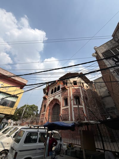 A woman stands in front of a white van, holding a woven fan. She is wearing a red top and a blue scarf. In the background, there is an old, ornate building with red and white detailing, likely a heritage site in Delhi, India. The building has arched windows and a large arched entrance. Overhead, numerous power lines crisscross the sky, obscuring the blue sky and white clouds. Several other white vans are parked in front of the building. A metal fence topped with barbed wire surrounds a portion of the property. To the left, a modern yellow building with glass windows stands next to the heritage structure. The scene appears to be a street in a busy urban area.