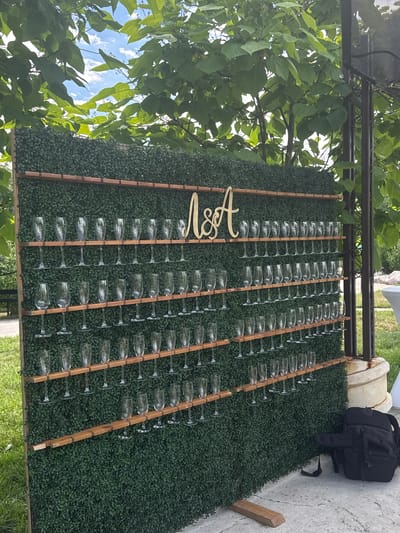The image shows an outdoor event setup with a wall covered in green artificial foliage. Wooden shelves are attached to the foliage wall, holding numerous champagne glasses. A golden monogram, "M&A", is mounted on the top shelf. To the right of the foliage wall, a black backpack is visible on the ground, and a pillar with decorative metalwork extends upwards. The scene appears to be a celebration or wedding, likely in Panchevo, Bulgaria, given the context provided, though the specific location details cannot be visually confirmed from the image alone. The background features lush green trees and a glimpse of a cloudy sky.