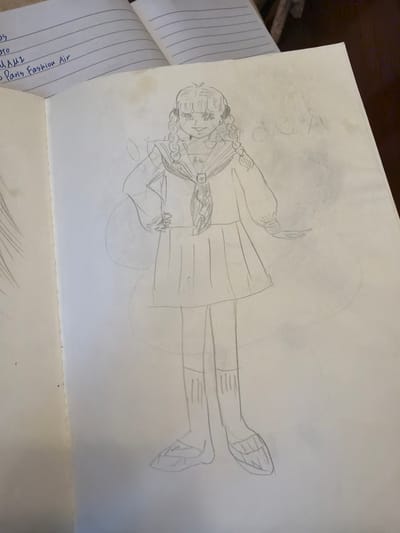 The image shows a pencil sketch of a girl in a school uniform on a white sheet of paper. The sketch is somewhat faded and surrounded by a rough outline of a circle. The girl is depicted wearing a pleated skirt, a top with a scarf, knee-high socks, and shoes. Her hair is styled with braids or twists adorned with circular elements. The sketch is located on top of a lined notebook, which is open. The notebook contains handwriting, where some of the words are discernible, including "Paris Fashion Air" and "MAMI."