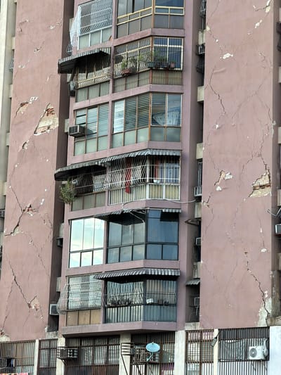 The image shows the exterior of a multi-story apartment building in Maracay, Venezuela, exhibiting significant structural damage. The building's facade is a muted rose color and displays large, prominent cracks that expose underlying brickwork in several areas, suggesting a potential structural compromise.
The building has multiple windows, many of which are protected by metal bars or grates. Some windows have awnings installed above them. Several air conditioning units are visible mounted on the exterior walls. Small potted plants are present on some of the balconies.
The lower levels of the building also feature large, barred windows or openings. A satellite dish is visible attached to one of the lower sections. The overall impression is one of disrepair and potential instability. No people are visible in the image. The environment appears to be urban, with other buildings or structures implied to be nearby.