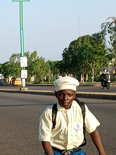 In Garko, Nigeria, on a clear day, a young boy wearing a yellow shirt, blue jeans, and a white beret is seen in the foreground. He has a black backpack on his shoulders and a belt with a decorative buckle. The boy is facing the camera, looking directly at it. Behind him, a street is visible with sparse traffic. A yellow tricycle and a motorcycle with a rider are in motion on the road. A tall green pole with white signs is on the left side of the street. Trees line the background of the scene.
