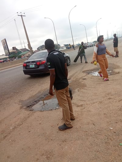 A medium shot shows a group of people on a dusty, unpaved road next to a multi-lane highway. A dark gray Hyundai Elantra is parked on the highway in the background. A man wearing a black t-shirt and brown pants stands in the foreground, facing away from the camera. To his right, a woman in tan pants and a blue top walks with a yellow object in her hand. To her left, a man in a green shirt carries a long, flat object. In the distance, more people and vehicles are visible. The sky is overcast. The scene appears to be in Kagini, Nigeria, indicated by street signs in the background and the overall environment.
