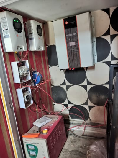 The image depicts an indoor electrical installation in Mowe, Nigeria, featuring solar energy components. On the wall, three white inverter units are mounted, two smaller ones on the left and a larger "Felicitysolar" branded unit on the right. Below the smaller inverters are two open electrical junction boxes with visible wiring. A blue electrical component is connected to wiring on the wall. A "Century Automatic Voltage Stabilizer" sits on the floor. Red and black wires snake across the floor, connecting to the electrical equipment. The wall on the left has red and white stripes. The wall on the right features a black and white circular pattern.