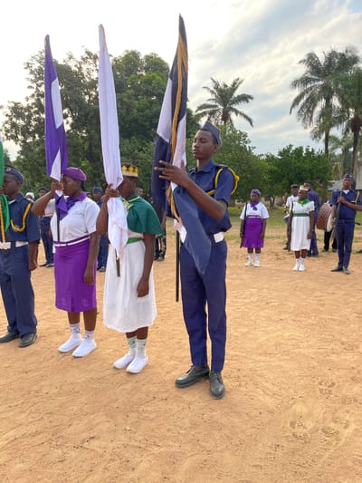 A group of people are arranged outdoors on a sandy surface, likely in Tabu, Nigeria, based on the user’s instructions. Some individuals hold flags of different colors - white, purple and white, and white, black, and gold. The people holding the flags wear various uniforms: a dark blue uniform with a matching cap, a white dress with a green cape and golden crown, and a white shirt and purple skirt with a purple beret. Other people are visible further back in the scene, some wearing similar dark blue uniforms and another white dress. Palm trees and lush green vegetation are visible in the background, under a partly cloudy sky.