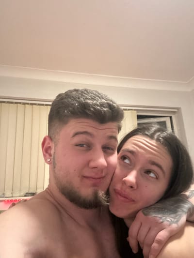 The scene depicts two individuals, an adult male and an adult female, in close physical proximity within a private indoor residence in Corby, United Kingdom. The male, presenting with short dark curly hair, faded sides, a light stubble beard, and a stud earring in his left ear, is shirtless and looks directly at the camera with a slight smile. The female, characterized by long dark hair and a visible tattoo sleeve on her left arm, rests her head against the male's right shoulder/chest area. She is looking upwards and to her right, away from the camera, with a slightly pensive expression. Her left arm is positioned around the male's torso. The background consists of a plain light-colored wall. A window with closed vertical blinds is partially visible behind the male to the left, and another window is visible behind the female to the right. A strip of red ambient lighting is observed along the lower-left edge of the frame. The ceiling is light-colored with decorative coving visible at the top.