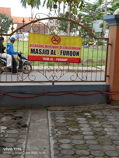 A sign in Indonesian reads "DILARANG MEROKOK DI LINGKUNGAN MASJID AL - FURQON", which translates to "Smoking is prohibited in the area of MASJID AL - FURQON", located in Kecamatan Haurgeulis, Indonesia (latitude: -6.46, longitude: 107.943). The sign is mounted on a black metal gate with a decorative scroll design. A red and white "no smoking" symbol is visible on the sign. Two people are riding a motorcycle in the background. The motorcycle is blue with a black seat and a red frame. The pavement in the foreground is made of large, irregularly shaped gray cobblestones. There is a red electrical wire running along the base of a concrete wall in the foreground. The scene is set in an outdoor area with green grass and trees in the background.
