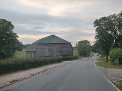 The image shows a rural road in Kirchnüchel, Germany, with a large wooden barn on the left side of the road. The barn is weathered and has a dark green roof. There are two square windows on the upper part of the barn wall. A dense green hedge runs along the front of the barn, separating it from the road. The road curves to the right, and several cars are visible driving away from the viewer. The surrounding landscape is green with trees on both sides of the road. The sky is overcast with a soft, diffused light, suggesting it might be early morning or late evening. There is a large stone on the right side of the road, near the trees.