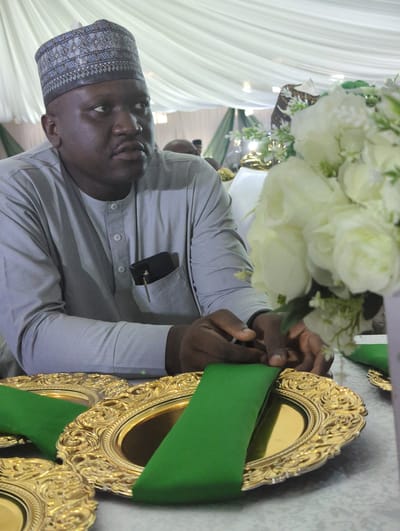 The image depicts a medium shot of a man wearing a grey traditional outfit and a patterned cap. He is seated at a table adorned with golden ornate plates and green napkins. A bouquet of white flowers is visible to his right. A mobile phone and a key are tucked into his chest pocket. His hands are resting on the table in front of him. The background consists of draped white fabric, suggesting an indoor event setting, possibly in Unguwar Rimi, Nigeria.