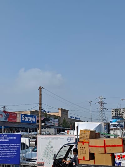 The image depicts a daytime street scene within a commercial district in Eshanguzar, Uzbekistan, under a predominantly clear blue sky with a single cumulus cloud visible on the left.
In the foreground, a dark-colored three-wheeled cargo vehicle, resembling a motorized rickshaw, is positioned slightly right of center. A male driver, wearing a dark cap, is visible in the driver's seat, looking forward. The vehicle's cargo bed is heavily loaded with multiple cardboard boxes of various sizes, secured with red and white strapping tape. Some boxes bear labels with text and barcodes. To the left of the vehicle, a large informational board displays a map-like diagram, possibly a site plan, accompanied by Cyrillic text. Further left, a blue billboard with white Cyrillic text, partially obscured by a metal fence, is visible. Behind the fence, several white passenger cars are parked.
The midground features a row of light-colored commercial buildings with arched architectural details. Visible signage on these buildings includes "Saroyli" (blue text on white), "SAVDO MARKAZI" (Cyrillic for "Trade Center"), and "КАМСКАЯ ПОСУДА" (Cyrillic for "Kamskaya Tableware"). Multiple utility poles and overhead power lines crisscross this section of the scene.
In the background, a large metal lattice electrical transmission tower stands prominently on the right. Beyond the tower, a multi-story building under construction with exposed concrete framework is visible, alongside another building featuring a distinct blue-domed roof. The overall scene suggests active commercial operations and urban infrastructure.
