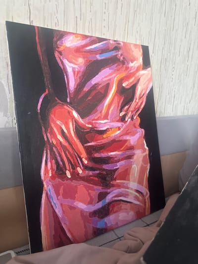 A painting on a canvas sits on a surface, likely a table or desk. The painting features an abstract rendering of two figures, primarily rendered in shades of red, pink, purple, and orange, against a black background. The figures' forms are not clearly defined but seem to depict bodies with visible hands. The surface underneath the painting appears to be textured and is partially obscured by other objects, including a dark-colored object and fabric. The location Высокий, Russia is not visually evident in the image.