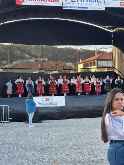Here is a detailed description of the image:

**Overview:** The image shows a scene at an event in Barcelos, Portugal. A group of performers in traditional folk costumes are on a stage with sponsor banners displayed prominently. A person is taking a selfie with the stage in the background. The environment is a paved area, suggesting an outdoor public space.

**People:**
*   **Performers:** Approximately 10-12 individuals are dressed in traditional Portuguese folk costumes. Their outfits feature red skirts, white shirts, and black hats.
*   **Selfie-taker:** A woman is positioned in the foreground, facing the camera, taking a selfie. She wears a white top and blue jeans.
*   **Other Person:** Standing in front of the stage on the right, facing to the stage.

**Objects:**
*   **Stage:** A stage is constructed, possibly for a performance, with a black backdrop.
*   **Banners:** Sponsor banners for "Miranda Supermercados" and "Ferrail" (a railway company) are visible on the left side of the stage.
*   **Barros & Barbosa Banner:** Another banner with "Barros & Barbosa Mat. de construção, Lda" is displayed on the stage.
*   **Paved Area:** The foreground consists of a paved area, suggesting a plaza or street.
*   **Buildings:** In the distance, residential buildings and houses.

**Actions and Interactions:**
*   **Performing:** The group is on stage, and it seems as though they may be in the middle of a performance or about to begin.
*   **Taking Selfie:** The woman in the foreground is taking a photo of herself with the performance in the background.

**Environment:**
*   **Outdoor:** The event takes place outdoors, likely in a public space like a town square or plaza.
*   **Barcelos, Portugal:** Given the traditional costumes and the presence of Portuguese company banners, the location is presumed to be in Barcelos, Portugal.