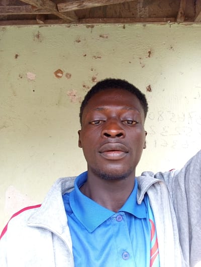 A portrait-style selfie features a dark-skinned young man looking directly at the camera. He has short, dark hair and a slight goatee. He is wearing a blue collared shirt with two visible buttons and a gray hooded jacket with red trim. The background consists of a pale green, textured wall with patches of peeling paint and some faint, illegible writing. Above the wall, wooden beams are visible, suggesting an outdoor or partially enclosed structure. The lighting is somewhat harsh, creating shadows on the man's face. The image appears to be taken outdoors, possibly in Gindin Dutse, Nigeria, given the context provided.