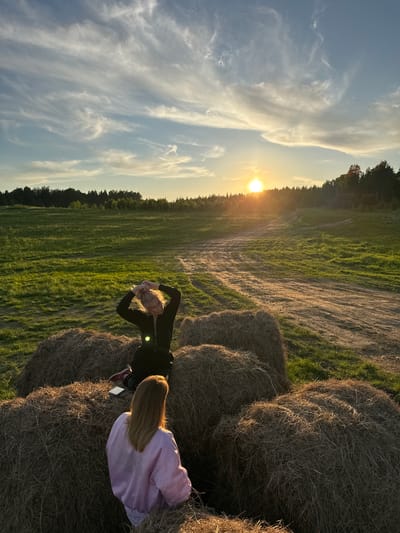 The image captures a serene outdoor scene at sunset in what appears to be a rural or agricultural setting, possibly in Стеклянный, Russia. The warm, golden light of the setting sun bathes the landscape, creating long shadows and highlighting textures.

In the foreground and midground, two individuals are seated on large, round hay bales. One person, positioned further from the camera, has long blonde hair and is wearing a dark long-sleeved top. They have their arms raised and clasped behind their head, looking towards the setting sun, possibly stretching or enjoying the view. The second person, closer to the camera and seen from behind, also has long blonde hair and is wearing a light purple jacket. A white smartphone or similar device rests on the hay bale next to them.

The hay bales are prominently featured, providing a rustic seating area. Beyond the bales, a vast, green grassy field stretches into the distance, traversed by light dirt paths. A dense line of trees forms the horizon, silhouetted against the bright sky where the sun is low.

The sky is a pale blue, adorned with wispy, elongated cirrus clouds that catch the golden light from the sun, creating a dynamic and picturesque backdrop. The overall mood is peaceful and contemplative, suggesting a moment of relaxation at the end of the day. There is no visible text in the image.