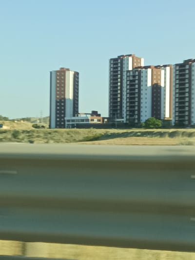 The media file shows a photograph of several tall apartment buildings against a clear blue sky. The buildings have a modern design with alternating sections of white and brown/red cladding, and rows of dark-colored windows. The buildings appear to be under construction or recently completed, as some windows are missing or incomplete. The scene is set in a semi-arid landscape with dry, grassy hills and some scattered trees. In the foreground, a dark barrier, possibly a highway sound barrier or median, runs horizontally across the image. The lighting suggests it is daytime, likely late afternoon or early morning, given the long shadows cast by the buildings. There are no people visible in the image. The setting appears to be a developing area, possibly on the outskirts of a town or city. No text is visible in the media file. The location is identified as Yahşihan, Turkey.