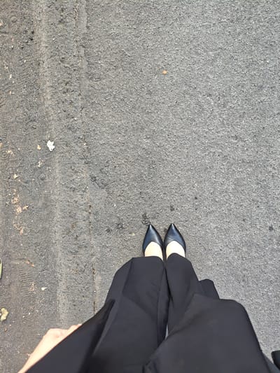 A first-person perspective shot shows a person wearing black pointed-toe flats and black pants standing on a paved surface. The paved surface appears to be asphalt, with some small stones and debris scattered on it. There is a slight discoloration or seam running vertically up the center of the frame. The lighting suggests it is daytime. The location is not identifiable as Sapporo, Japan based on the visual information provided.