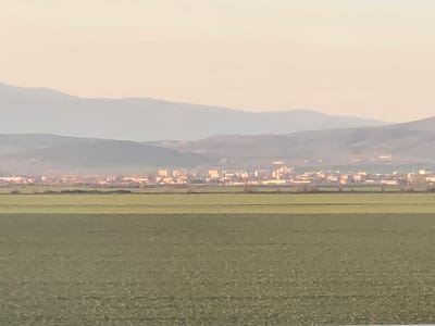 The image displays a wide-angle, distant view of the city of Diadovo, Bulgaria. In the foreground, a vast, flat expanse of cultivated land, uniformly covered with green, low-lying vegetation (likely crops), occupies the lower half of the frame. A faint, dark horizontal line, possibly a distant treeline or dense scrub, separates the foreground field from the urban settlement. The mid-distance presents the city of Diadovo, characterized by numerous architectural structures, including multi-story buildings, some of which exhibit bright reflections consistent with direct, low-angle sunlight. These structures are distributed horizontally across the visible cityscape. The background is composed of multiple layers of rolling hills and mountains, appearing muted and indistinct due to atmospheric haze. The sky above is a pale, featureless light tone. No individuals, specific actions, or interactions are discernible.