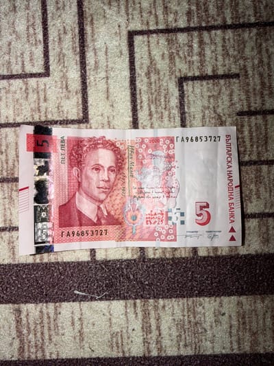 A Bulgarian five leva banknote lies on a patterned carpet. The banknote features a portrait of Ivan Milev, a Bulgarian artist who lived from 1897 to 1927. The banknote is marked with the serial number ГА96853727 and issued by the Bulgarian National Bank (БЪЛГАРСКА НАРОДНА БАНКА). The denomination "5" is prominently displayed.
