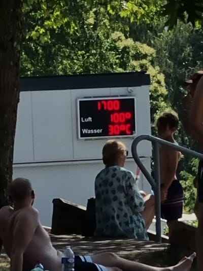The image depicts an outdoor scene, likely at a swimming pool or waterfront in Saarbrücken, Germany, on a sunny day. Several individuals are present, some partially visible. In the foreground, a person is lying down, their back to the camera. Further back, another person is seated, looking towards a digital display. A young person, also shirtless, is partially visible behind a metal railing.
The focal point of the image is a digital display mounted on a white structure. The display shows three lines of text and numbers in red. The top line reads "1700". The middle line displays "100°". The bottom line shows "30° C". Below these readings, there are two labels: "Luft" and "Wasser", indicating that the display shows the air temperature and water temperature, respectively.
The scene appears relaxed and summery, with lush green trees in the background suggesting a natural or park-like setting. The presence of people in swim attire or shirtless implies a warm weather activity, possibly swimming or sunbathing. The overall mood seems to be one of leisure and enjoyment of the outdoors.