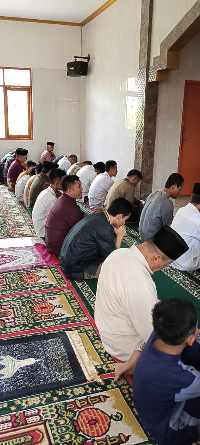 The image shows a group of men in a mosque located in Kecamatan Kutawaringin, Indonesia. They are sitting on prayer rugs with their heads bowed in prayer. The men are wearing traditional clothing and some have black skullcaps on their heads. The room is clean and well-lit, with white walls and a tiled floor. There is a window on the left side of the room that shows trees and a building in the distance. A speaker is mounted on the wall in the top left corner of the room. There is a door on the right side of the room that leads to another room. The men are engaged in silent prayer, their heads bowed in unison, and the atmosphere is calm and serene. There is a young boy sitting on the floor, with his back to the camera, watching the men praying.  The mosque is decorated with traditional Islamic patterns and designs.
