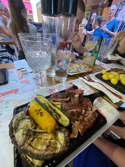 The scene depicts a table in a restaurant likely in Bologna, Italy, as indicated by the "Vicolo Ranocchi, 4/c - Te" address on the placemat, with the word "BOLOGNA" visible.
**People:**
Several people are seated around the table, some identifiable by their torsos, arms, or heads. There are individuals wearing blue shirts, while others are partially visible in the background.
**Subjects:**
The main subject is food presented on several serving dishes.
- A black rectangular dish contains sliced beef.
- Grilled vegetables, including eggplant, zucchini, and yellow pepper, are also present on the black dish.
- Another dish contains cooked potatoes.
- A small dish filled with small white shells and nuts
**Objects:**
- Several glasses, some filled with liquid, are on the table.
- A metal fork is present on the table.
- A smartphone is also visible on the table.
- A decorative photo frame.
- Paper placemats with a design.
**Actions:**
People appear to be in the act of dining and socializing.
**Environment:**
The environment is a restaurant with a wooden table. The scene is well-lit.