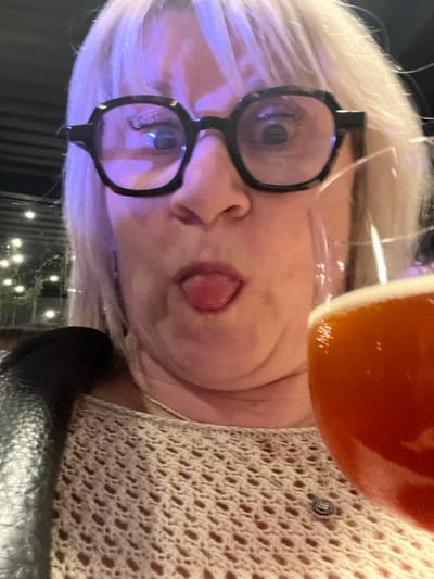 A woman with short white hair and large, round glasses poses at an angle. She wears a light-colored, crocheted top and a thin necklace. She holds a glass with an orange liquid in front of her face. The background shows outdoor lights and greenery. The image appears to be taken at night with dim lighting.