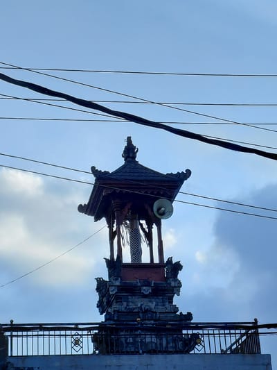 A traditional Balinese temple structure stands against a cloudy blue sky, with several black power lines crossing the frame. The temple features multiple tiers, with intricate stone carvings and a dark, multi-layered roof. A white speaker is mounted on the side of the upper section of the temple. A black metal railing with decorative elements runs along the base of the structure. The image appears to be taken in the city of Sukasada, Indonesia, given the architectural style and context. There are no people visible in the image.